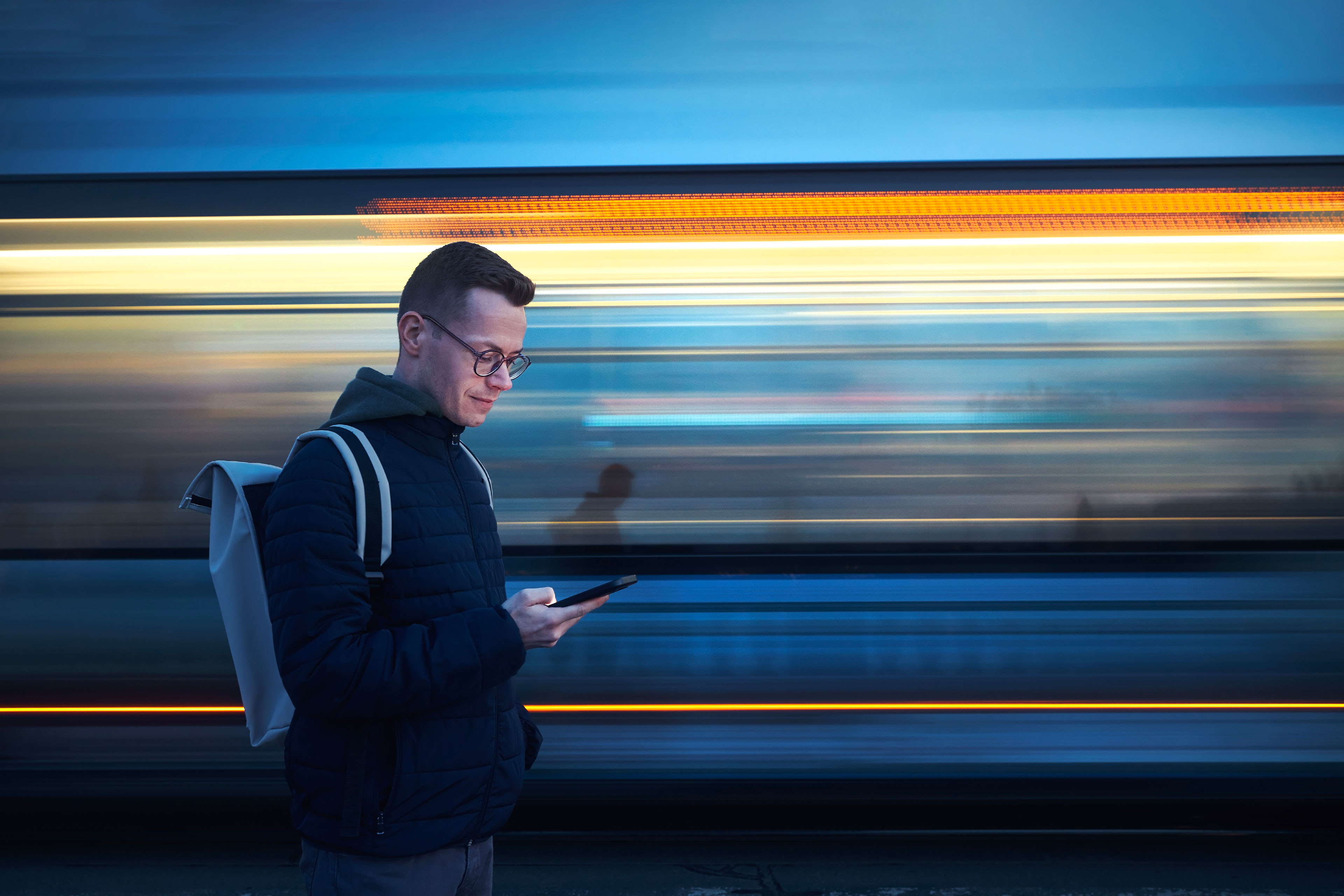 Man with backpack holding and using phone against tram in blurred motion