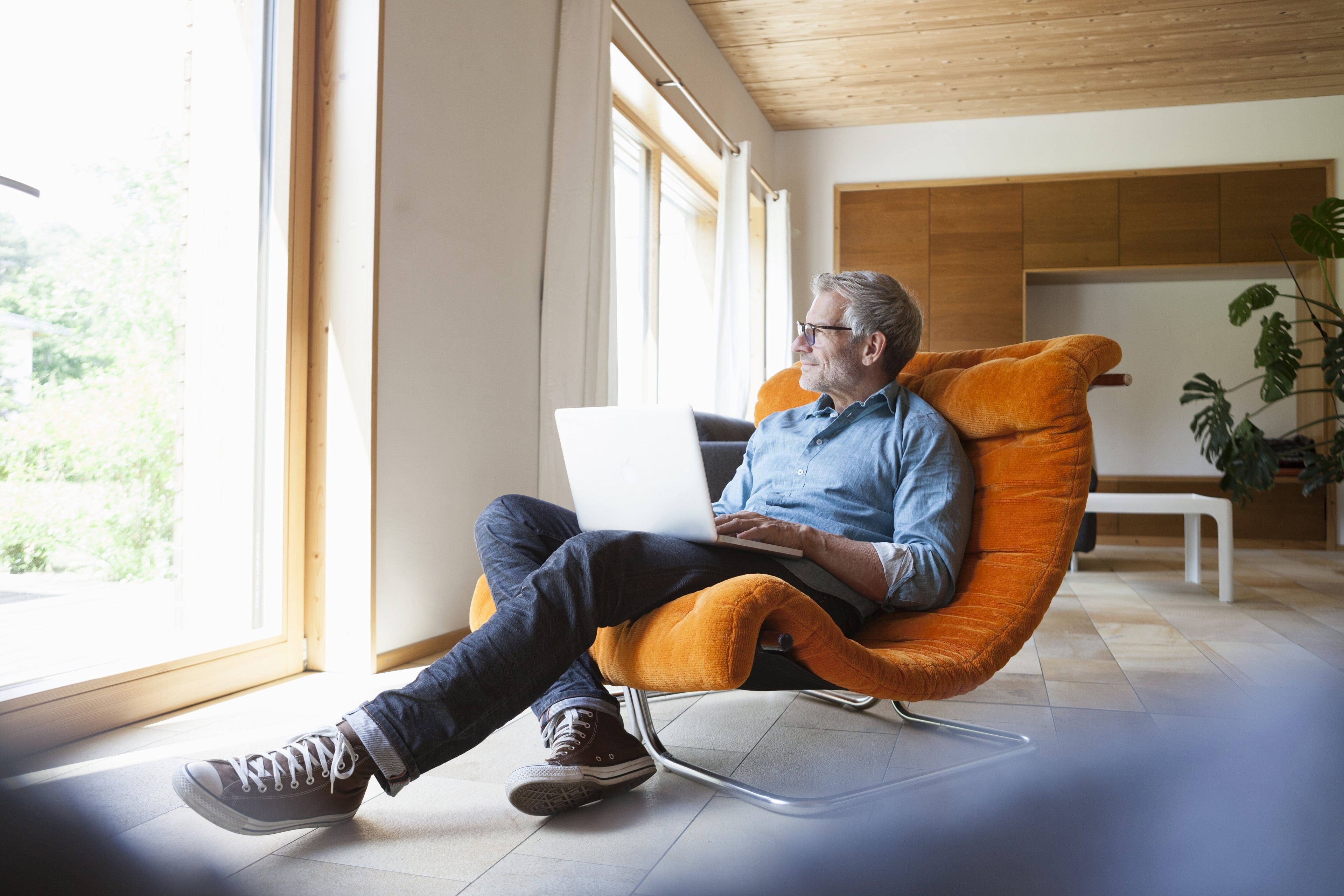 A man sitting on a chair in a hall and looking outside through the window with a laptop on his lap