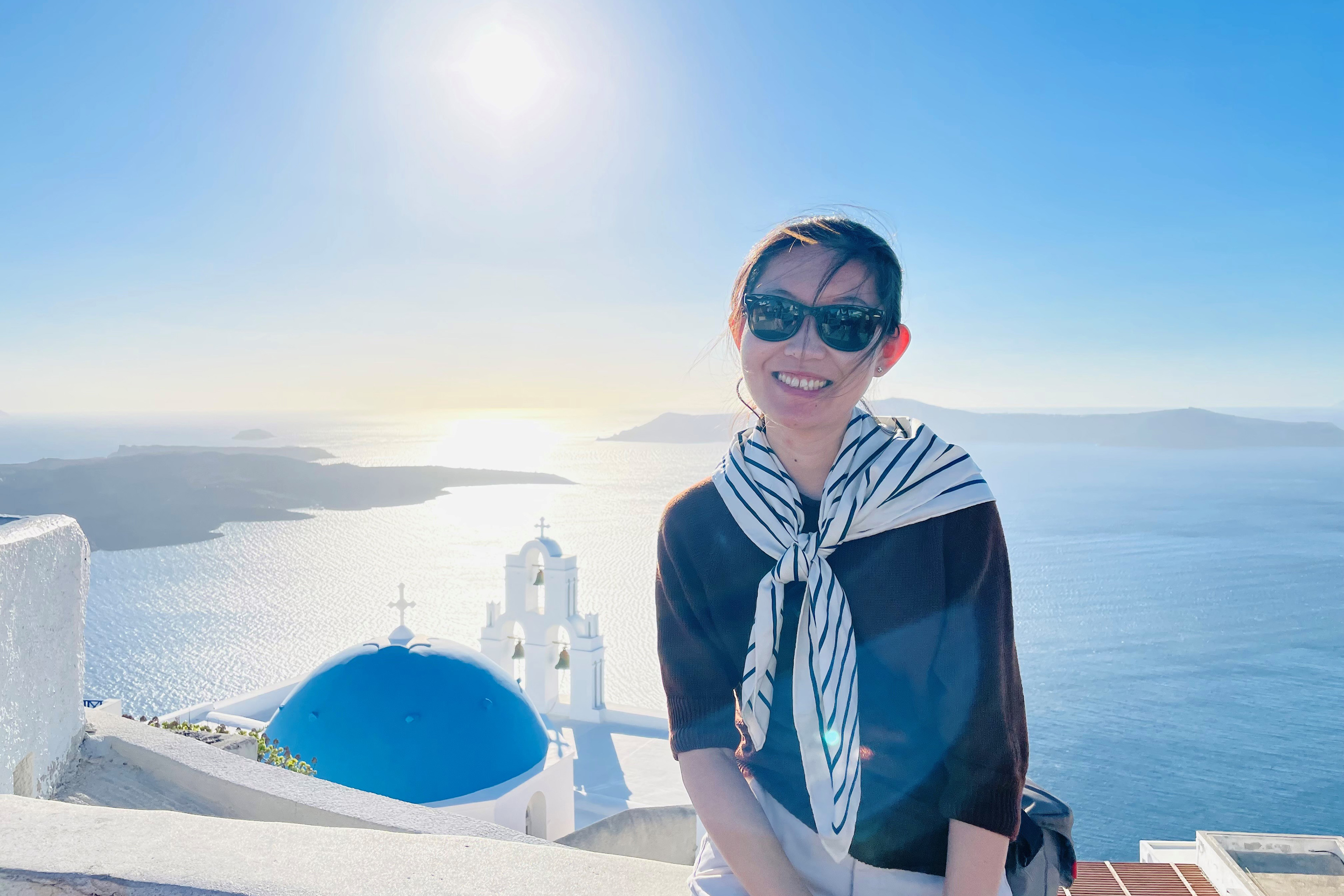 A woman is smiling and wearing glasses with the ocean in the background.