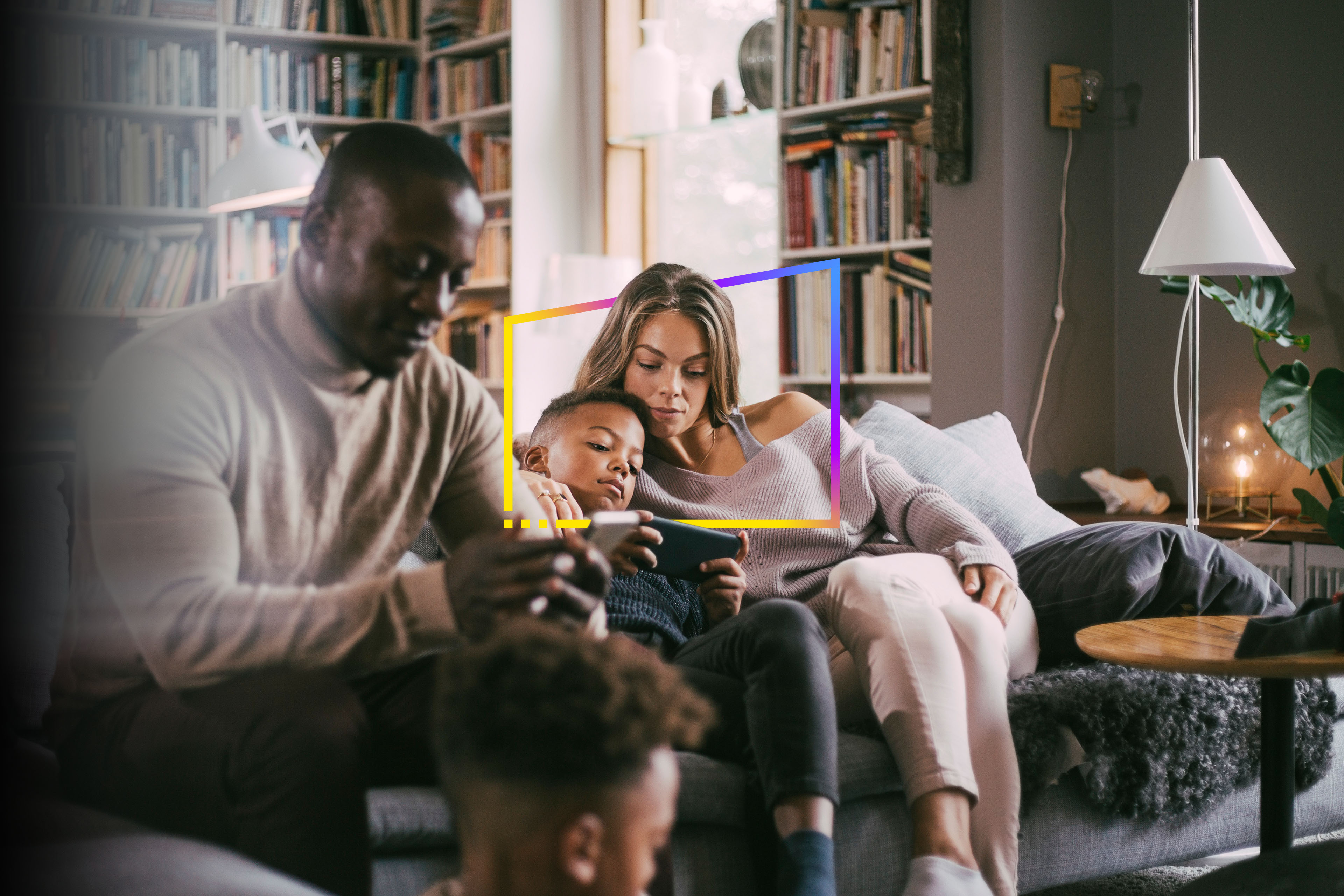 A family sits on a couch in a cozy living room with a bookshelf, floor lamp, and natural light from a window.