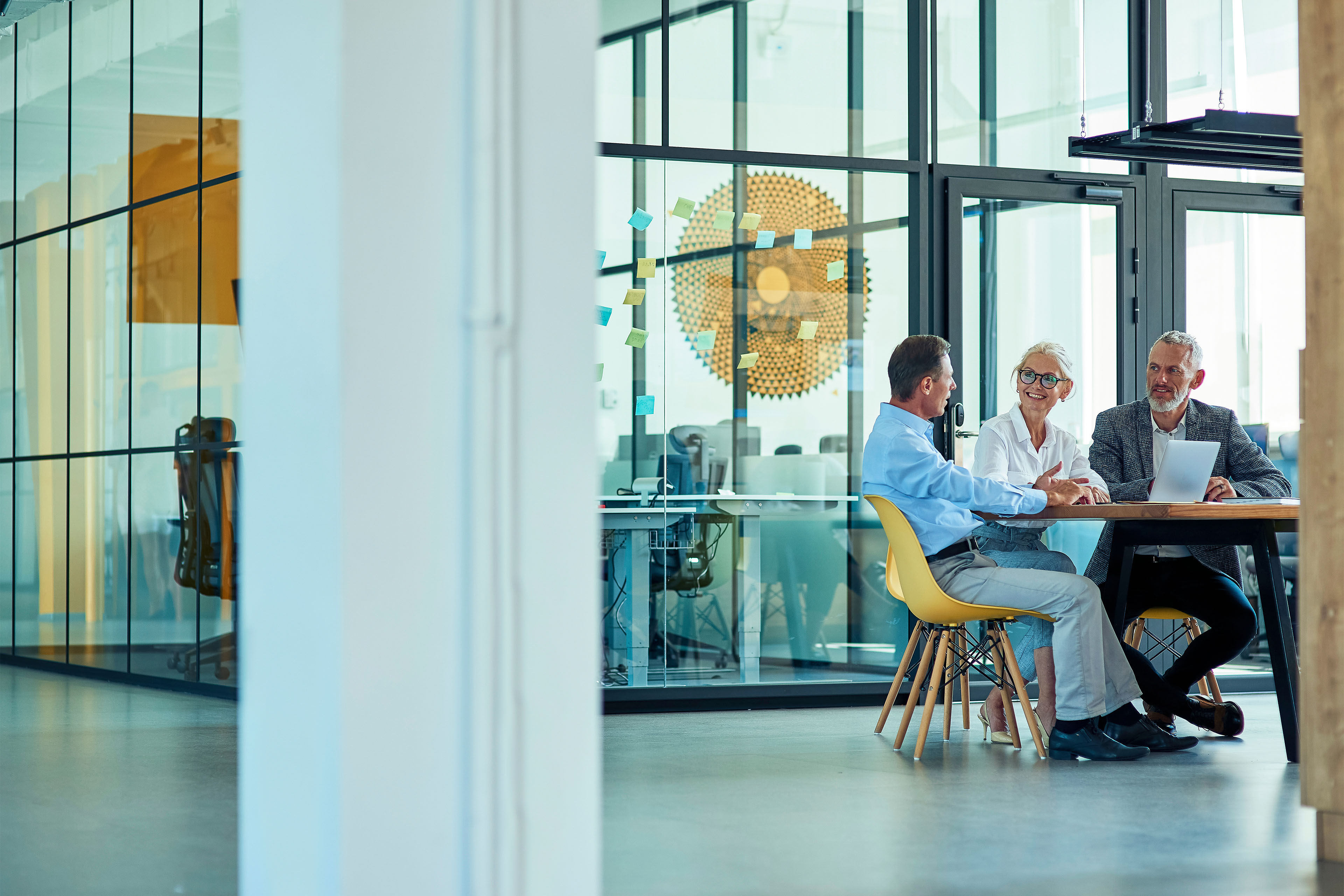 Kolme kypsää kollegaa istuu modernissa toimistossa tai coworking-tilassa, käyttää kannettavaa tietokonetta ja