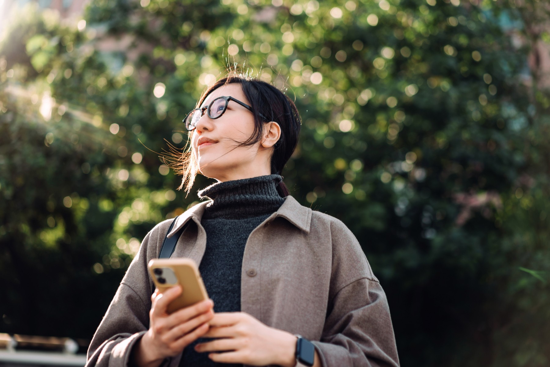 Low-angle-portrait-of-young-asian-woman-using-smartphone-in-park-against-sunlight.