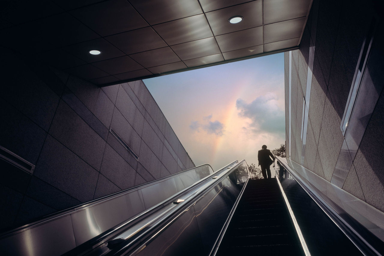 Escalator of subway station with businessman moving towards dramatic sky with rainbow.